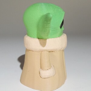 May include: A small, green and beige figurine of a character from the Star Wars franchise. The character has large ears and a robe.