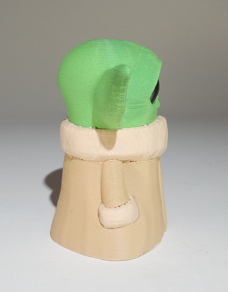 May include: A small, green and beige figurine of a character from the Star Wars franchise. The character has large ears and a robe.