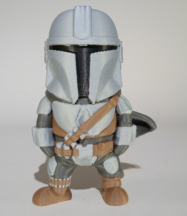 K&ouml;nnte beinhalten: Eine 3D-gedruckte Figur eines Mandalorianer-Charakters mit silbernem Helm und R&uuml;stung, einer braunen Brustplatte und einem passenden G&uuml;rtel. Die Figur hat einen kleinen Umhang und steht aufrecht.