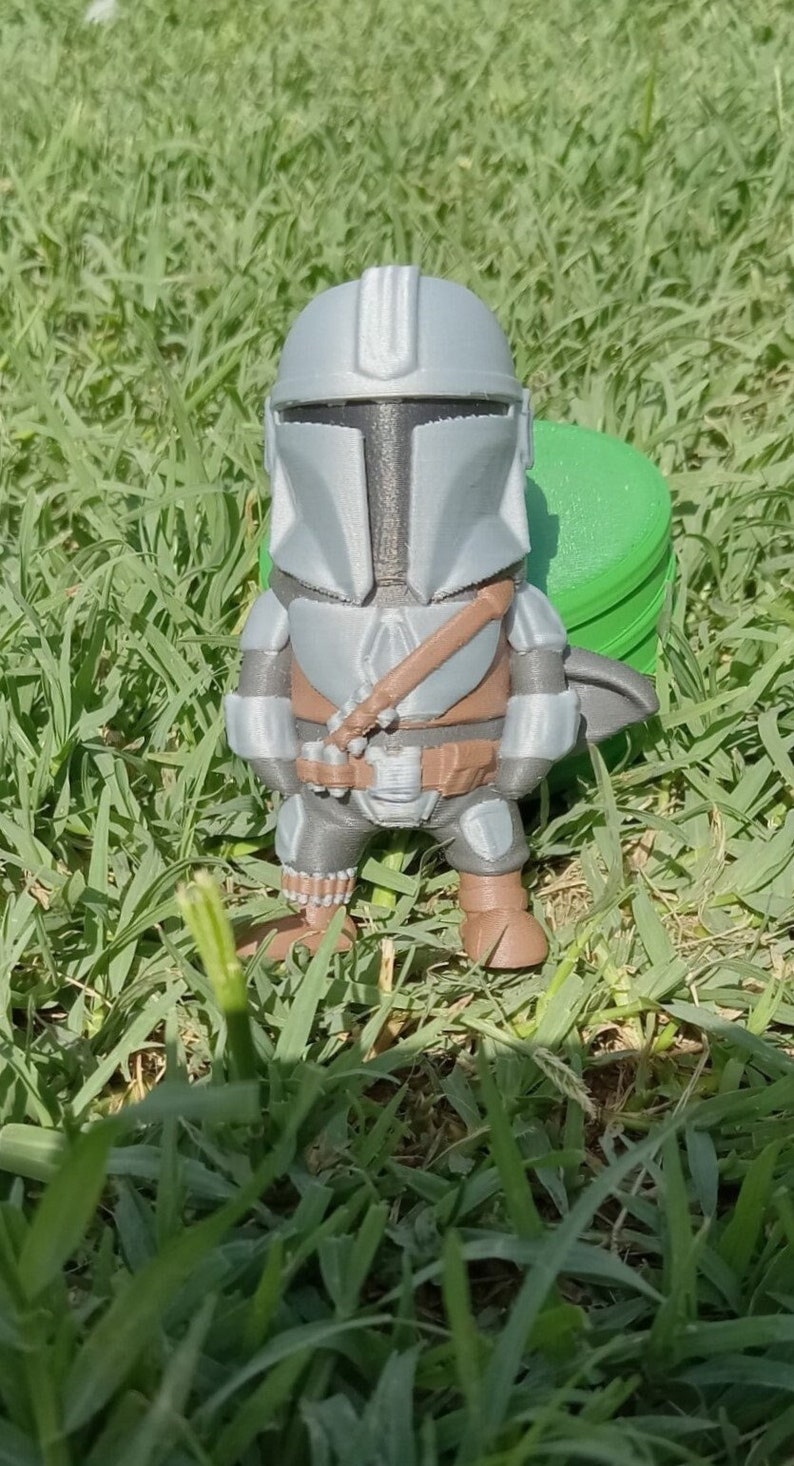 K&ouml;nnte beinhalten: Eine 3D-gedruckte Mandalorianer-Figur im Chibi-Stil. Die Figur ist haupts&auml;chlich grau mit braunen und schwarzen Akzenten, einschlie&szlig;lich Helm, R&uuml;stung und Blaster. Sie steht auf einer gr&uuml;nen Grasfl&auml;che.