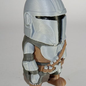 K&ouml;nnte beinhalten: Eine 3D-gedruckte Figur eines Mandalorianers mit silbernem Helm und R&uuml;stung sowie braunen und grauen Akzenten. Die Figur hat ein stilisiertes Design und steht aufrecht.