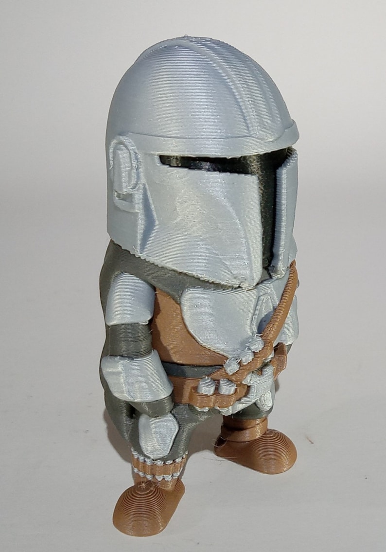 K&ouml;nnte beinhalten: Eine 3D-gedruckte Figur eines Mandalorianers mit silbernem Helm und R&uuml;stung sowie braunen und grauen Akzenten. Die Figur hat ein stilisiertes Design und steht aufrecht.
