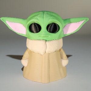 May include: A green and pink 3D printed figurine of a character from the Star Wars franchise. The character has large ears, big eyes, and is wearing a tan robe.