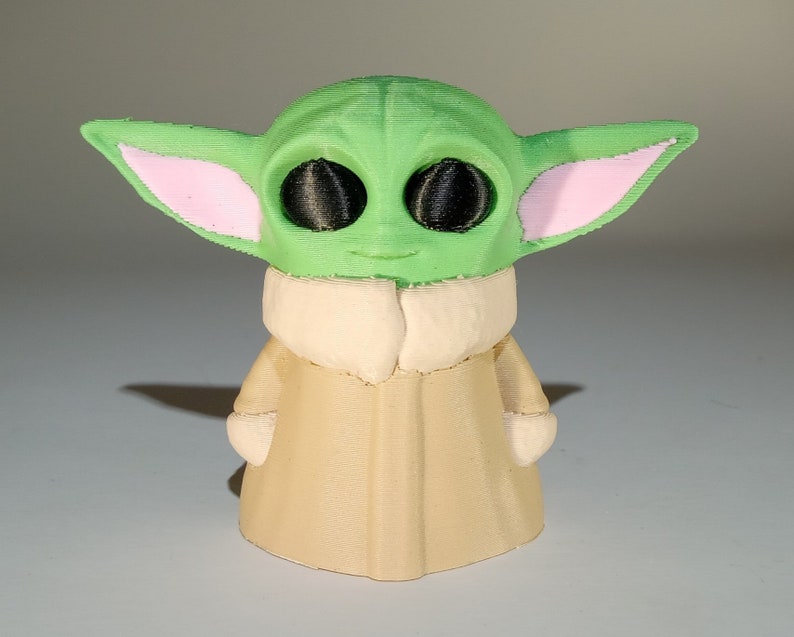 May include: A green and pink 3D printed figurine of a character from the Star Wars franchise. The character has large ears, big eyes, and is wearing a tan robe.