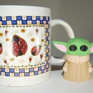 May include: A white ceramic mug with a blue and white checkered pattern and a ladybug design. The mug has a handle and is sitting on a white surface. A small green and pink figurine is in front of the mug.