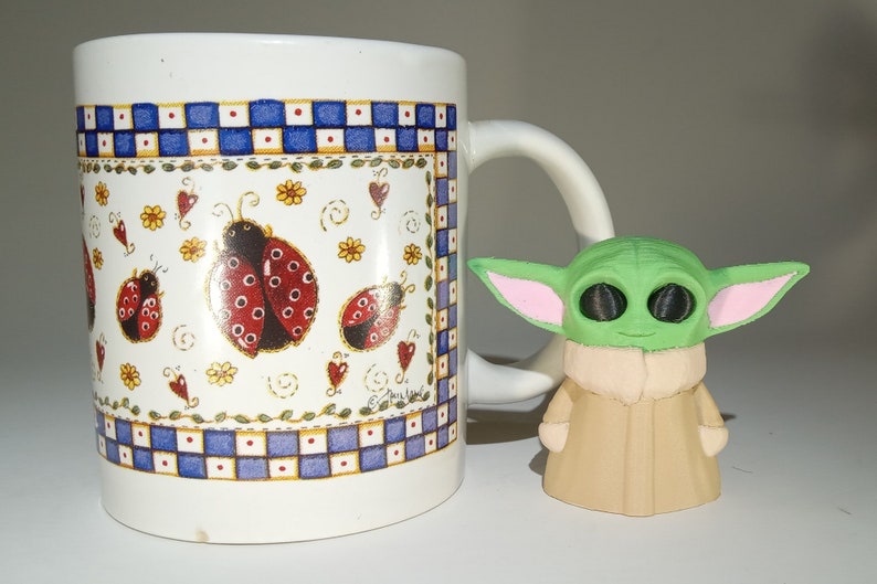 May include: A white ceramic mug with a blue and white checkered pattern and a ladybug design. The mug has a handle and is sitting on a white surface. A small green and pink figurine is in front of the mug.