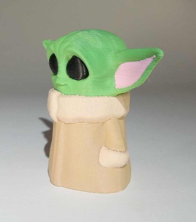 May include: A green and beige 3D printed figurine of a character from the Star Wars franchise. The character has large ears and big eyes.