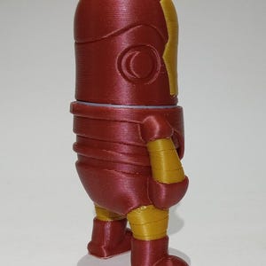 May include: A red and gold 3D printed figurine of a superhero. The figurine is standing with its arms at its sides and its legs slightly apart. The figurine has a red head and torso, with a gold face mask and gold accents on the arms and legs.