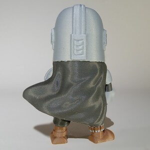 K&ouml;nnte beinhalten: 3D-gedruckte Figur einer Figur aus einer Science-Fiction-Serie, von hinten gesehen. Die Figur hat einen silbernen Helm, einen dunkelgrauen Umhang und braune Stiefel. Die Figur steht auf einer wei&szlig;en Oberfl&auml;che.