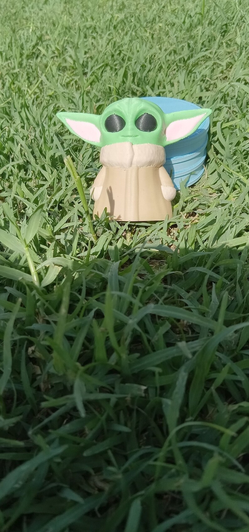 May include: A green and brown figurine of a character from the Star Wars franchise, known as "The Child" or "Baby Yoda", standing on a blue and white circular object. The figurine is in a grassy area.