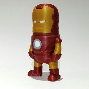 May include: A red and gold 3D printed figurine of Iron Man. The figurine is standing with its arms at its sides and has a serious expression on its face.