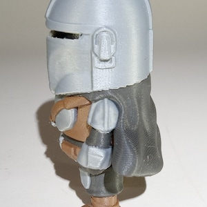 K&ouml;nnte beinhalten: 3D-gedruckte Figur einer Figur aus einer Science-Fiction-Serie. Die Figur hat einen silbernen Helm, eine braune R&uuml;stung und einen dunkelgrauen Umhang. Die Figur steht aufrecht und hat ein detailliertes, strukturiertes Aussehen.