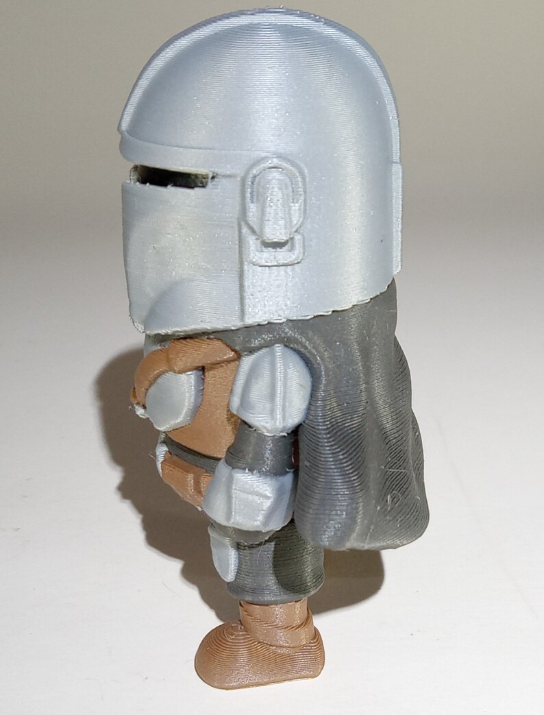 K&ouml;nnte beinhalten: 3D-gedruckte Figur einer Figur aus einer Science-Fiction-Serie. Die Figur hat einen silbernen Helm, eine braune R&uuml;stung und einen dunkelgrauen Umhang. Die Figur steht aufrecht und hat ein detailliertes, strukturiertes Aussehen.
