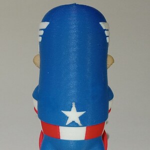 May include: A red, white, and blue 3D printed figurine of Captain America. The figurine is standing with its arms at its sides and is wearing a red, white, and blue costume. The figurine is wearing a blue helmet with a white star on the front. The figurine is standing on a red base.