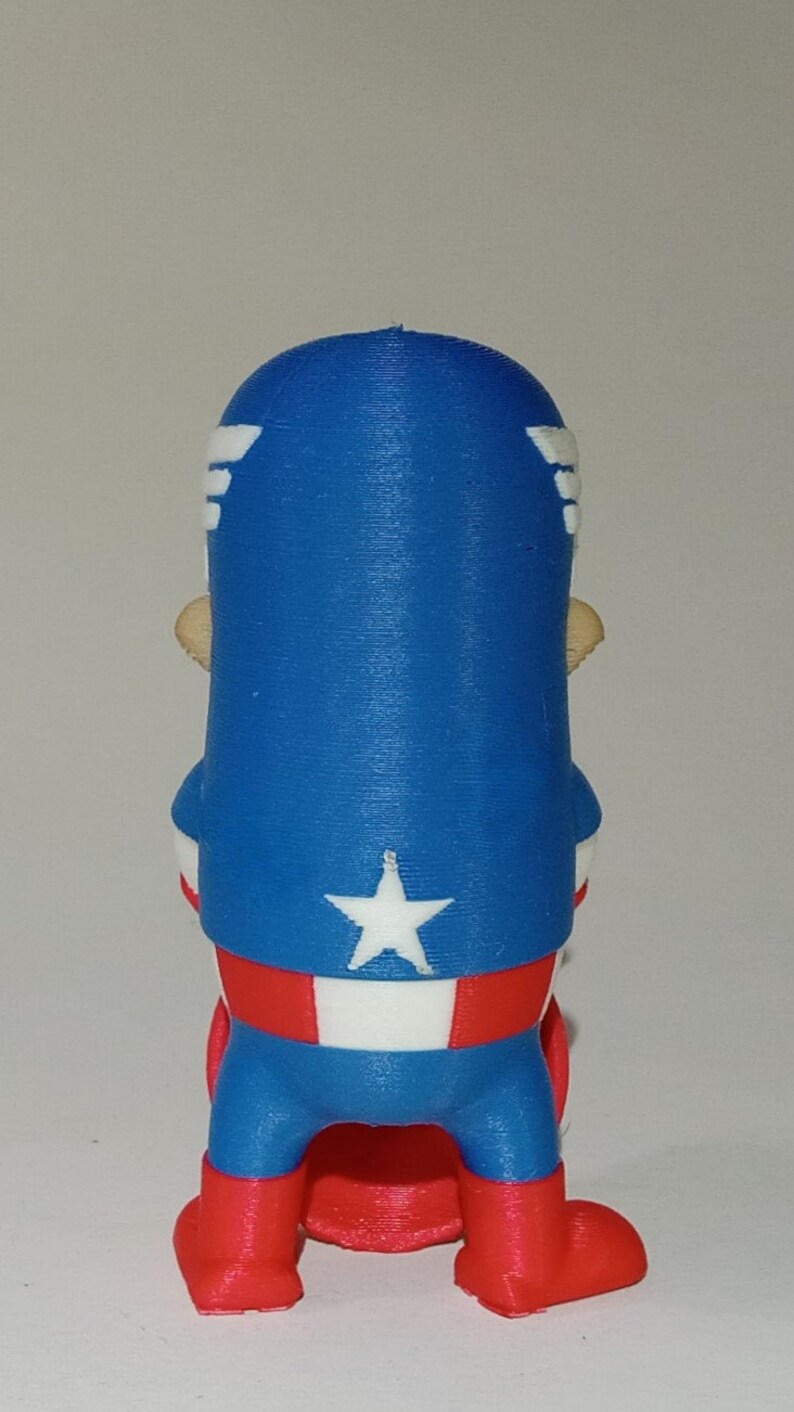 May include: A red, white, and blue 3D printed figurine of Captain America. The figurine is standing with its arms at its sides and is wearing a red, white, and blue costume. The figurine is wearing a blue helmet with a white star on the front. The figurine is standing on a red base.