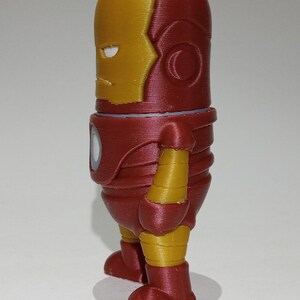 May include: A red and gold 3D printed figurine of Iron Man, a superhero from Marvel Comics. The figurine is standing with its arms at its sides and its head tilted slightly to the side. The figurine is about 6 inches tall.