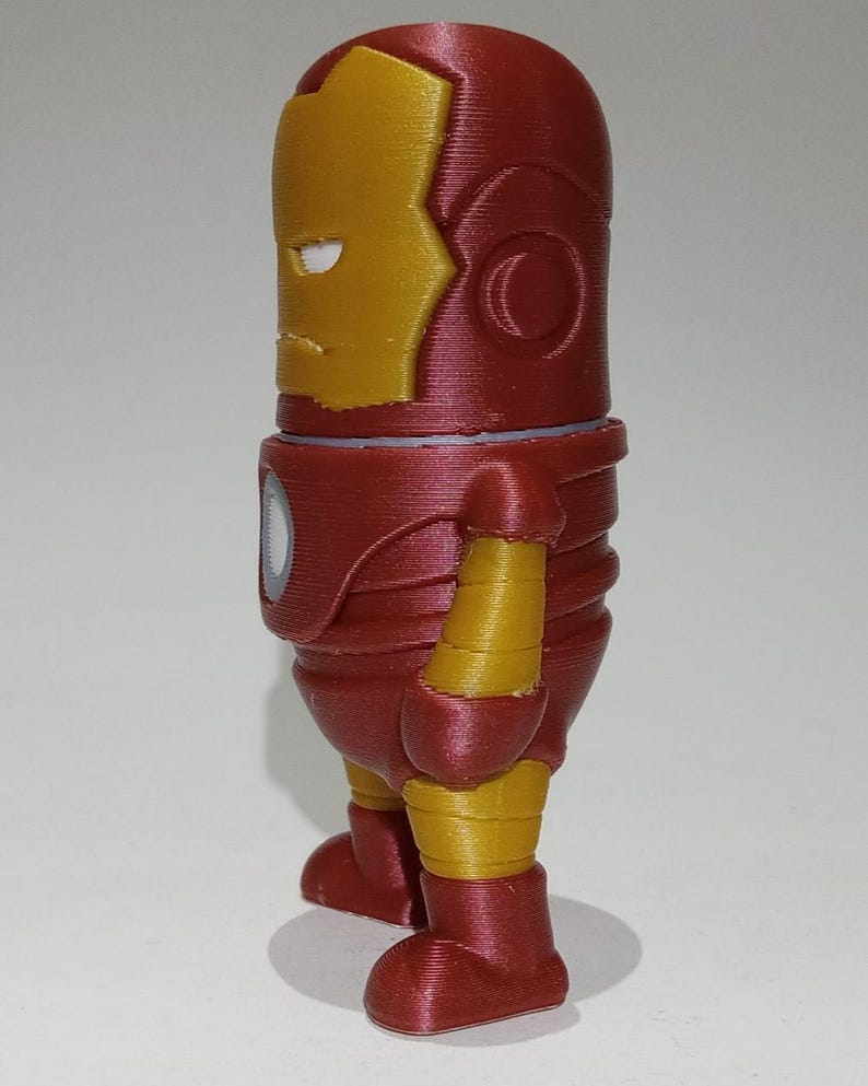 May include: A red and gold 3D printed figurine of Iron Man, a superhero from Marvel Comics. The figurine is standing with its arms at its sides and its head tilted slightly to the side. The figurine is about 6 inches tall.