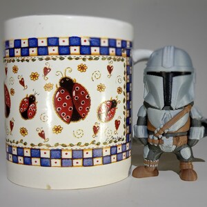 K&ouml;nnte beinhalten: Eine wei&szlig;e Keramiktasse mit einem blau-wei&szlig;en Karomuster und Marienk&auml;fer-Illustrationen. Daneben steht eine grau-braune Mandalorianer-Actionfigur, ein Sammlerst&uuml;ck aus dem Star Wars-Franchise.