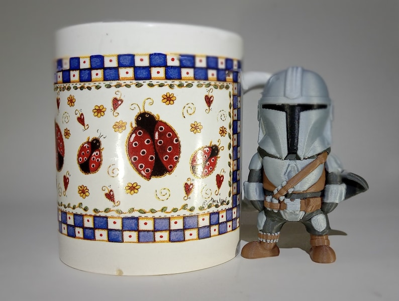 K&ouml;nnte beinhalten: Eine wei&szlig;e Keramiktasse mit einem blau-wei&szlig;en Karomuster und Marienk&auml;fer-Illustrationen. Daneben steht eine grau-braune Mandalorianer-Actionfigur, ein Sammlerst&uuml;ck aus dem Star Wars-Franchise.