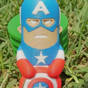 May include: A small, 3D printed figurine of Captain America. The figurine is blue, red, and white and features a classic Captain America design with a star on his chest and a shield in his hands. The figurine is standing on a green grassy surface.