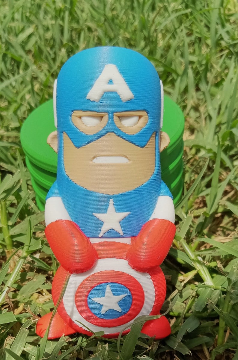 May include: A small, 3D printed figurine of Captain America. The figurine is blue, red, and white and features a classic Captain America design with a star on his chest and a shield in his hands. The figurine is standing on a green grassy surface.