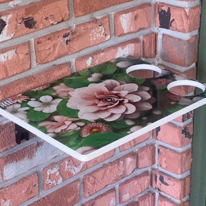 Puede incluir: Estante con estampado floral con borde blanco, montado en una pared de ladrillo. El estante presenta una gran flor rosa rodeada de flores blancas y rosas más pequeñas y hojas verdes. Se ven dos recortes circulares.