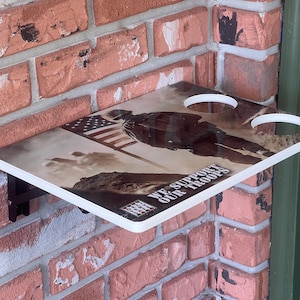 Pode incluir: Prateleira retangular de parede com um design patriótico. A prateleira apresenta uma imagem de um soldado e da bandeira americana, com o texto "WE SUPPORT OUR TROOPS". Possui dois recortes circulares e uma borda branca.