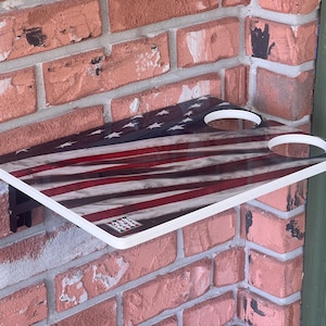 May include: A rectangular shelf with a white border, featuring a distressed American flag design in red, white, and blue. The shelf has two circular cutouts and is mounted on a brick wall.