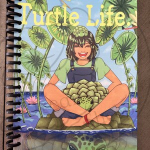 May include: A spiral bound notebook with a colorful illustration of a girl sitting on a turtle. The notebook cover features the text "Turtle Life" in yellow letters and the phrase "Slow, Steady, Success" in yellow letters at the bottom. The illustration features a blue background with green lily pads and a dragonfly.