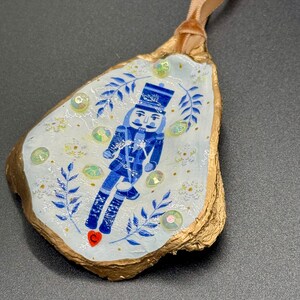 May include: A hand-painted oyster shell Christmas ornament featuring a blue nutcracker soldier design. The shell has a gold-painted edge and is adorned with iridescent sequins and a tan ribbon for hanging. The background is white with blue leaf accents.