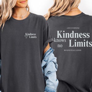 May include: Dark gray t-shirt with the text "Kindness knows no Limits" on the front and back. The back of the shirt also includes the phrases "Love Everyone" and "Be Kind to All Kinds". The shirt has rolled-up sleeves.