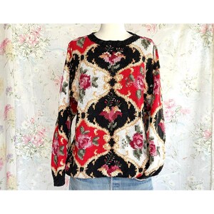 May include: A vintage sweater with a floral pattern. The sweater features a black, red, white, and gold design with roses. The sweater has a crew neck and long sleeves. The sweater is displayed on a mannequin.