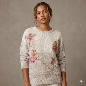 May include: A cream-colored sweater with a textured knit design. The sweater features floral embellishments in pink and peach tones, accented with gold-colored chain details. The sweater has a round neckline and long sleeves.