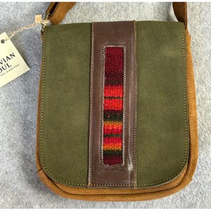 May include: Olive green crossbody bag with a brown leather strap and accents. The bag features a central vertical panel with a woven, colorful design in red, orange, and yellow. The tag reads "Bolivian Soul, Peru in every piece."