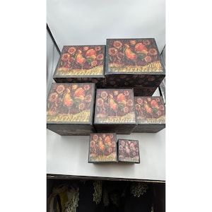 May include: A set of six square nesting boxes in varying sizes, each featuring a dark brown base with a printed design of a colorful rooster standing among sunflowers. The boxes have a consistent rustic aesthetic with matching patterns on all lids.