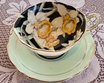 May include: A vintage teacup and saucer set with a black background and a floral design of white and yellow daffodils. The saucer is a light green color with a gold trim.