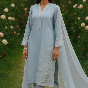 May include: A light blue, long-sleeved tunic and matching pants set with intricate silver embroidery. The tunic features a V-neck and a flowing, sheer scarf. The outfit is worn with silver earrings and a bracelet. The background is a garden with flowers.