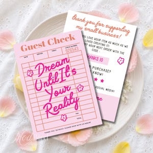Może przedstawiać: Dwie prostokątne papierowe karty z różowym i białym tłem. Górna karta ma wzór różowego rachunku z napisem "Dream Until It's Your Reality" wykonanym różową kursywą, małymi ikonami kwiatów oraz numerem kontrolnym 926543.
