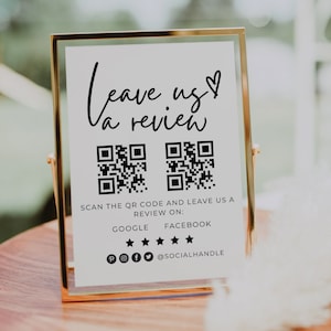 Peut inclure: Une pancarte blanche avec un cadre doré, affichant le texte "Leave us a review" avec un symbole de cœur. Deux codes QR sont affichés sous le texte, ainsi que des instructions pour scanner les codes et laisser un avis sur Google et Facebook. Des icônes de médias sociaux sont incluses en bas de la pancarte.