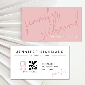 May include: A white business card with black text that reads "Jennifer Richmond Interior Designer". The card also includes a QR code, social media icons, and a website address. The text "Jennifer" is written in pink cursive at the bottom of the card.