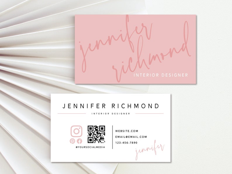May include: A white business card with black text that reads "Jennifer Richmond Interior Designer". The card also includes a QR code, social media icons, and a website address. The text "Jennifer" is written in pink cursive at the bottom of the card.