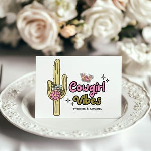 May include: A white rectangular card featuring a graphic of a light green cactus adorned with pink and blue flowers. Next to the cactus is a small butterfly, four-pointed stars, and the text Cowgirl Vibes T-SHIRTS & APPAREL in pink and olive green.