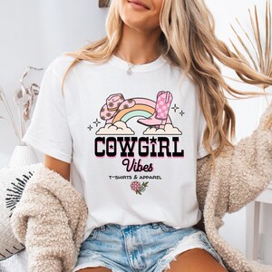 May include: A white t-shirt featuring a graphic print of a pink cowboy hat, a pink checkered cowboy boot, and a rainbow over clouds. The text reads COWGIRL Vibes T-SHIRTS & APPAREL, accompanied by a small floral illustration below the text.