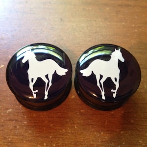 Może przedstawiać: Dwie czarne zatyczki do uszu z białym wzorem sylwetki konia. Zatyczki są okrągłe i mają błyszczące wykończenie. Koń jest przedstawiony w pozycji biegu, z wyciągniętymi nogami. Zatyczki leżą na drewnianej powierzchni.