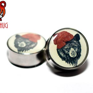 Könnte beinhalten: Paar schwarze und weiße Bären-Ohrmanschetten mit einer roten Mütze auf dem Bären. Die Manschetten sind aus Metall und haben eine glatte Oberfläche.