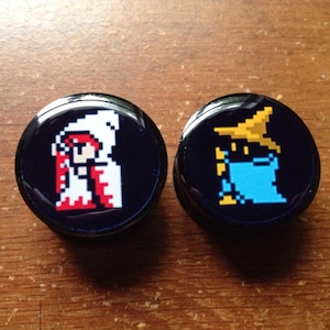 Puede incluir: Dos pendientes redondos negros con una imagen pixelada blanca y roja de un personaje femenino con una túnica blanca y una imagen pixelada azul y amarilla de un personaje masculino con una túnica azul y un sombrero amarillo.