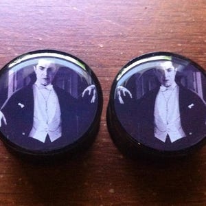 Może przedstawiać: Para czarnych rozszerzeń do uszu z okrągłą, czarną obwódką i czarno-białym obrazkiem mężczyzny w smokingu. Mężczyzna ma na sobie białą muszkę i poważny wyraz twarzy.