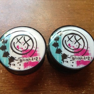 Puede incluir: Par de dilatadores de oreja negros y blancos con un gráfico azul, rosa y blanco del logotipo de Blink-182. El logotipo presenta una cara sonriente con una 'X' sobre cada ojo.