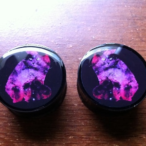 May include: A pair of black circular ear gauges with a galaxy print featuring a pink cat.