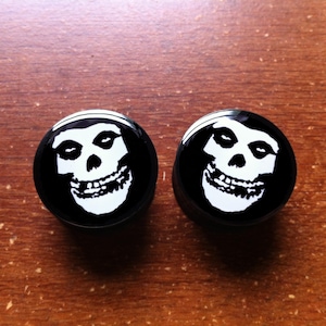 Puede incluir: Un par de expansores de oreja de calavera Misfits en blanco y negro. Los expansores son redondos y tienen una calavera blanca sobre un fondo negro.
